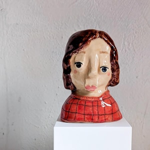 May include: A ceramic bust of a person with brown hair and blue eyes. The figure wears a red plaid shirt. The sculpture is displayed on a white pedestal against a white background.