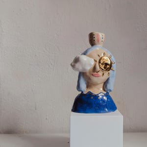 Könnte beinhalten: Eine Keramik-Büsten-Vase mit hellblauem Haar, einer weißen Wolke über dem rechten Auge und einem goldenen Sonnenemblem über dem linken Auge. Der Sockel ist dunkelblau mit goldenen Akzenten und eine kleine zylindrische Öffnung sitzt oben auf dem Kopf.