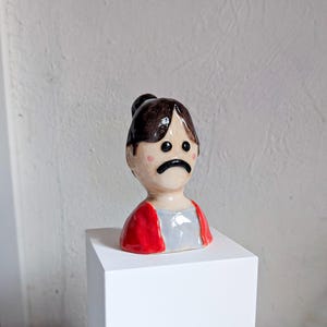 May include: A ceramic figurine of a person with a sad expression. The figure has brown hair in a bun, rosy cheeks, and is wearing a red jacket over a gray shirt. The figurine is displayed on a white pedestal.