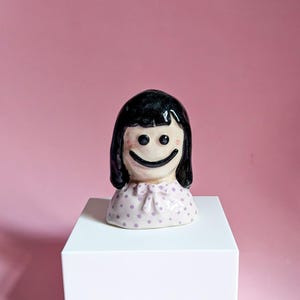 May include: A ceramic figurine of a smiling girl with black hair and a light pink dress with purple polka dots. The figurine is on a white pedestal against a pink background. The girl's face has a large, happy smile.