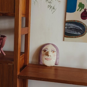 May include: A ceramic wall mask featuring a stylised human face with smooth, pale skin, light purple hair, brown eyes, and pink lips. The mask has a glossy finish and is positioned on a wooden shelf next to a wooden shelving unit and a wall painting.