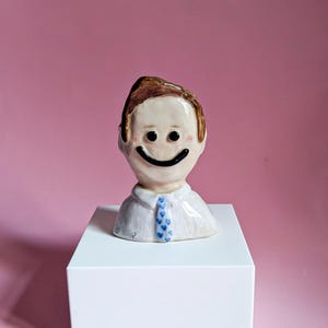 May include: A ceramic bust with a smiling face, brown hair, and a white shirt with a blue patterned tie. The sculpture is displayed on a white cube against a pink background. The face has black eyes and a black smile.