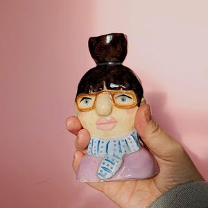 以下が含まれることがあります： ダークブラウンのお団子ヘア、オレンジ色の縁のメガネ、青い目、ピンクの唇をした頭の形の陶器製フィギュア。ラベンダー色の服の上に、ダークブルーのドット柄が入ったライトブルーのスカーフを巻いている。
