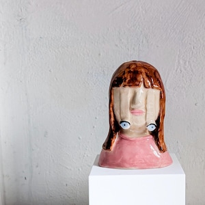 May include: A ceramic sculpture of a stylized face with brown hair and a pink top. The face has blue eyes and a unique design with elongated features. The sculpture is displayed on a white pedestal against a neutral background.