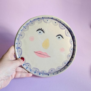 May include: A hand-painted ceramic plate with a smiling face design. The plate has a cream base with a blue scalloped border. The face features black eyelashes, pink cheeks, a yellow nose, and pink lips. A unique decorative piece for the home.