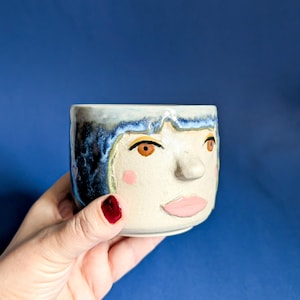 Könnte beinhalten: Eine Keramiktasse mit Gesichtsdesign. Die Tasse hat einen hellbeigen Sockel mit blau-weißem Rand und blauem Haar. Das Gesicht hat braune Augen, eine rosa Nase und Wangen sowie einen rosa Mund. Die Tasse wird von einer Hand mit rotem Nagellack gehalten.