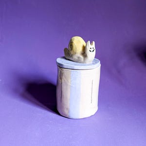 Può includere: Un vaso in ceramica con coperchio, con una figura di lumaca. Il vaso cilindrico è dipinto con strisce verticali beige, azzurro e giallo pallido. La lumaca è gialla con una faccia sorridente. Lo sfondo è viola.