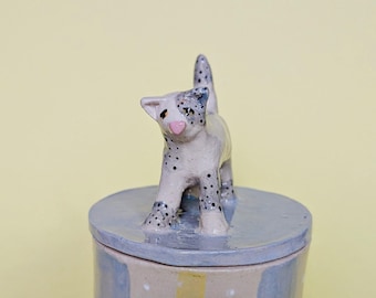 May include: A ceramic jar with a lid featuring a sculpted cat figurine on top. The cat is off-white with black spots and a pink nose. The jar body has vertical stripes in light blue, beige, and yellow with small white dots on the beige sections.