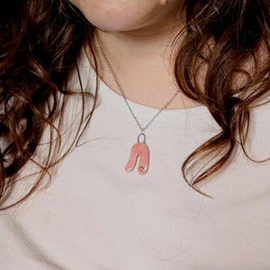 May include: A silver-toned chain necklace featuring a smooth, U-shaped pendant in a soft, muted pink hue. The pendant has a glossy finish and a small, circular indentation near the bottom right edge, suspended from a simple silver metal bail.
