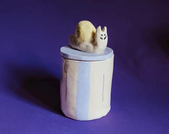 May include: A cylindrical ceramic jar with a light cream and pale blue vertical striped pattern. The lid features a sculpted snail figurine with a yellow shell and a cream-colored body, accented with black painted eyes and a small smiling mouth.