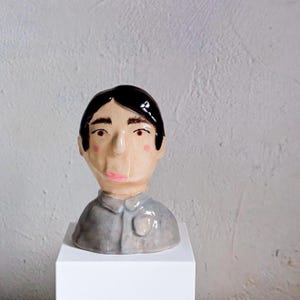 May include: A ceramic bust of a person with black hair, rosy cheeks, and pink lips. The figure wears a gray shirt with a badge. The sculpture is displayed on a white, square pedestal against a textured, off-white background.