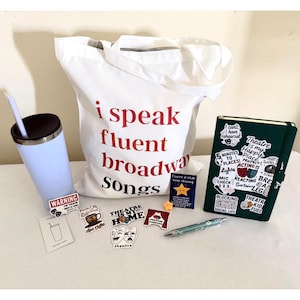 May include: A white canvas tote bag with the red and black text I speak fluent broadway Songs, a light blue tumbler with a straw, a dark green notebook covered in theatre-themed stickers, a pen, and several small decorative stickers and cards.