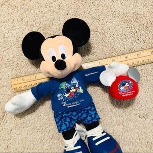 Puede incluir: Un juguete de peluche de Mickey Mouse con una camisa azul con el texto "Where magic lives." y pantalones de pijama azul y blanco. El juguete sostiene un sombrero de peluche de Mickey Mouse rojo con el mismo texto.