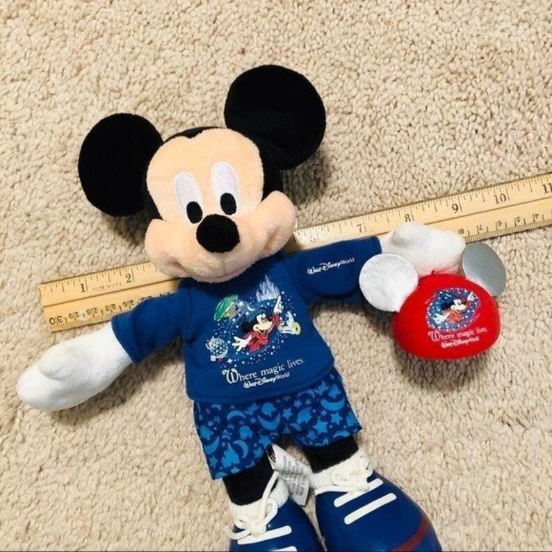 Puede incluir: Un juguete de peluche de Mickey Mouse con una camisa azul con el texto "Where magic lives." y pantalones de pijama azul y blanco. El juguete sostiene un sombrero de peluche de Mickey Mouse rojo con el mismo texto.