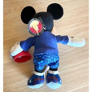 Puede incluir: Un juguete de peluche azul y rojo de Mickey Mouse que lleva una chaqueta azul y pantalones azules con un patr&oacute;n de estrellas y lunas. El juguete sostiene un sombrero rojo en su mano izquierda. El juguete tiene una etiqueta que dice "Disney Parks Authentic Original".