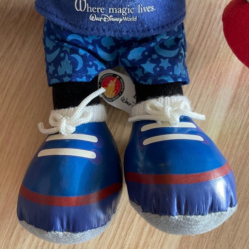 Puede incluir: Juguete de peluche azul y blanco con una camisa azul, pantalones con estampado azul y blanco y zapatillas azules con cordones blancos. El juguete tiene una etiqueta que dice "Where magic lives. Walt Disney World."