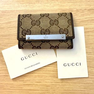May include: A brown and tan Gucci key case with the Gucci logo pattern. The key case has a silver metal bar with the Gucci logo. Two white cards with the Gucci logo are also present.