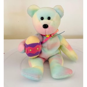 May include: A pastel tie-dye plush bear holding a purple and yellow Easter egg with pink and green accents.