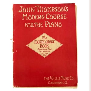 May include: A vintage red piano book titled "John Thompson's Modern Course For The Piano." The cover features white text and a diamond-shaped emblem that reads "The Fourth Grade Book." The book is from The Willis Music Co., Cincinnati, O.