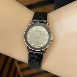Puede incluir: Un reloj de cuarzo ADOLFO dorado con una correa de cuero negro. La esfera del reloj es plateada con detalles de diamantes y agujas doradas. El reloj muestra la fecha y la palabra "Quartz".