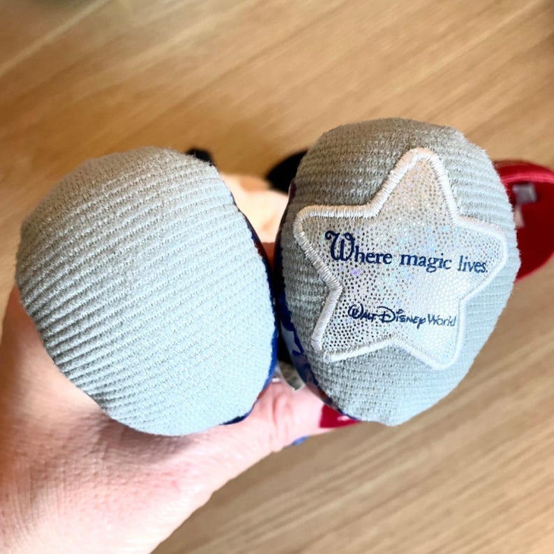 Puede incluir: Dos orejas de Mickey Mouse de felpa grises con un dise&ntilde;o de estrella azul y roja. La oreja derecha tiene una estrella blanca con el texto "Where magic lives" y "Walt Disney World" bordado en ella.