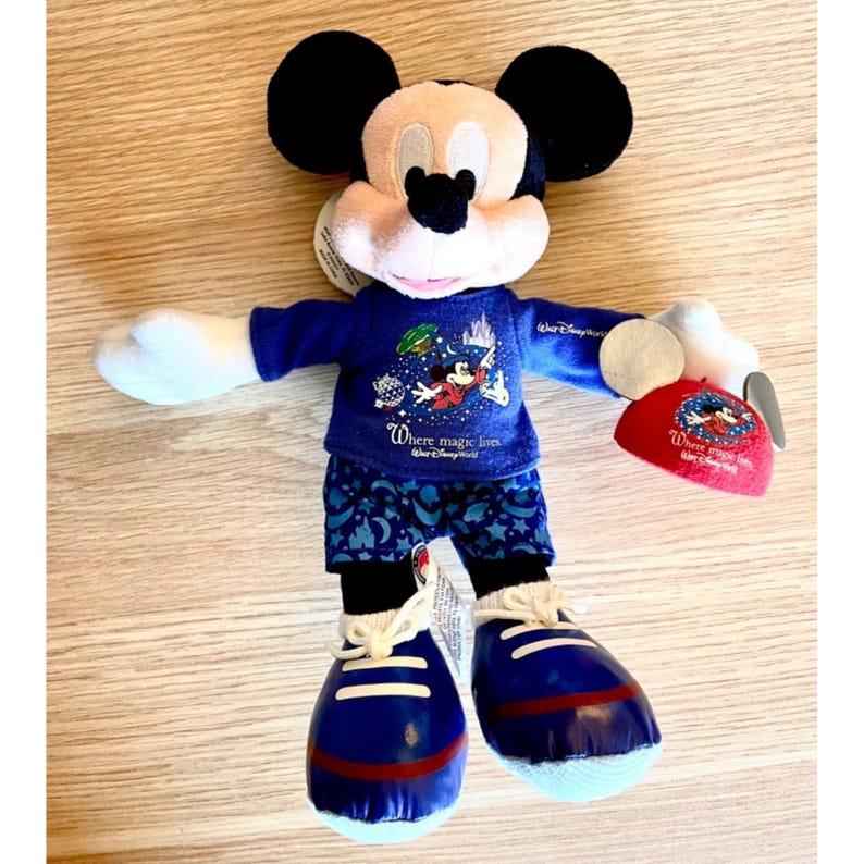 Puede incluir: Un Mickey Mouse de peluche azul y blanco que lleva una camisa azul con un gr&aacute;fico de Mickey Mouse y el texto "Where magic lives." Mickey Mouse lleva un pijama azul y blanco con un patr&oacute;n de estrellas. Mickey Mouse lleva zapatillas azules con cordones blancos y ribetes rojos. Mickey Mouse sostiene un sombrero rojo con un gr&aacute;fico de Mickey Mouse y el texto "Where magic lives."