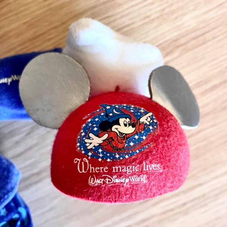 Puede incluir: Orejas rojas de Mickey Mouse con orejas plateadas y una parte superior blanca. Las orejas tienen un gr&aacute;fico de Mickey Mouse como mago con el texto "Where magic lives, Walt Disney World".