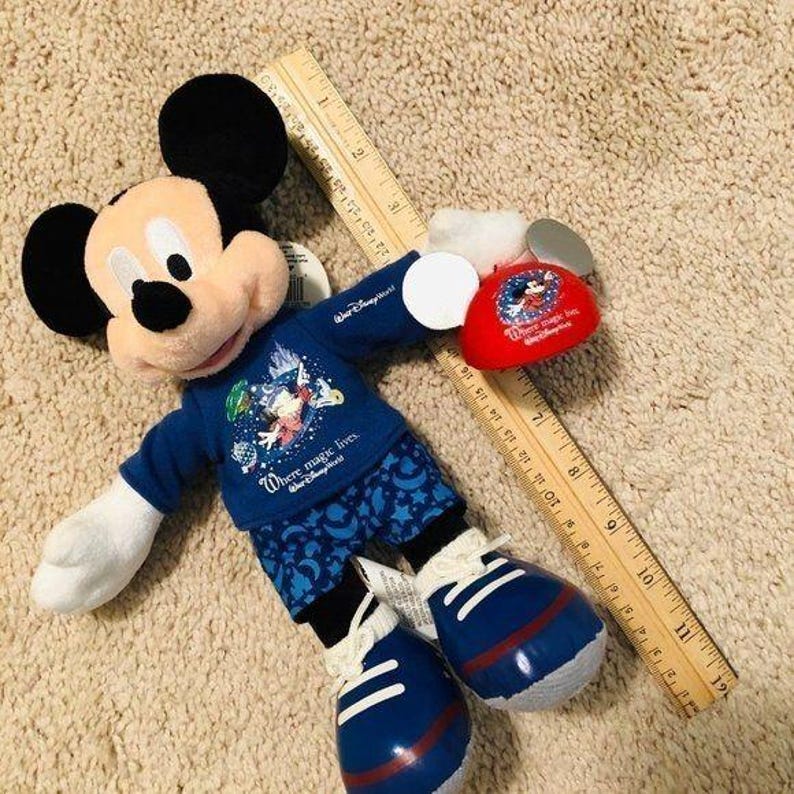 Puede incluir: Un juguete de peluche de Mickey Mouse con una camisa azul con el texto "Where magic lives" y pantalones azules con estrellas. El juguete sostiene una cabeza de peluche de Mickey Mouse roja y blanca con el texto "Where magic lives".