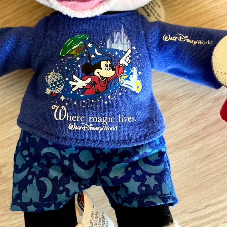 Puede incluir: Un juguete de peluche de Mickey Mouse que lleva una camisa azul con una ilustraci&oacute;n de Mickey Mouse y el texto "Where magic lives. Walt Disney World". La camisa tiene un patr&oacute;n de estrellas y medias lunas azules y blancas. El juguete lleva pantalones cortos azules con un patr&oacute;n similar de estrellas y medias lunas.