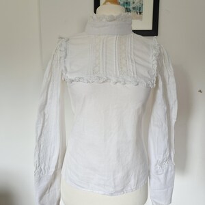 Op de afbeelding: Een witte blouse met lange mouwen, een hoge kraag, ingewikkelde kanten afwerking op het borstpaneel en ruches op de schouders. Het kledingstuk is voorzien van verticale plooien op het voorpand en nauwsluitende manchetten aan de mouwuiteinden.