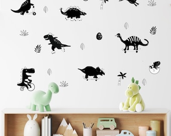 May include: Black and white dinosaur wall decals featuring a variety of dinosaurs riding skateboards, scooters, and bicycles. The decals are on a white wall with a wooden shelf below. The shelf has a variety of items on it, including a green stuffed dinosaur, a white ceramic bunny, and a wooden mountain range.
