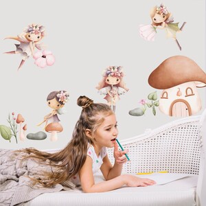May include: A young girl is sitting on a white couch with a blanket. She is looking at a wall with fairy and mushroom themed wall decals. The decals feature fairies with wings and flowers, mushrooms with houses, and green leaves.