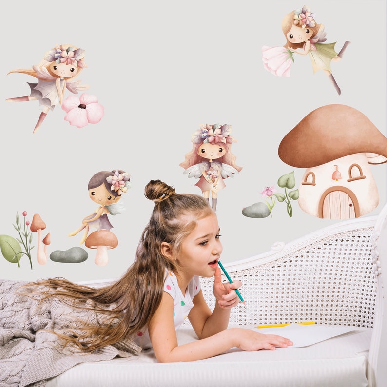 May include: A young girl is sitting on a white couch with a blanket. She is looking at a wall with fairy and mushroom themed wall decals. The decals feature fairies with wings and flowers, mushrooms with houses, and green leaves.