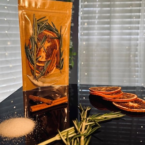 May include: A brown paper bag filled with dried orange slices, cinnamon sticks, and rosemary sprigs. Scattered around the bag are additional orange slices, cinnamon sticks, rosemary, and a pile of white granules, likely sugar or salt.