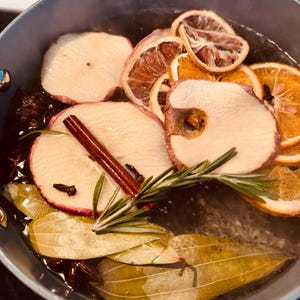 Może przedstawiać: Jasnoniebieski garnek wypełniony wodą i składnikami do gotowania. Widoczne są plasterki jabłek, pomarańczy i cytryn, a także laska cynamonu, gałązka rozmarynu, liście laurowe i anyż gwiazdkowaty. Składniki są w wodzie.