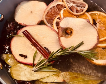 May include: A pot filled with water and various ingredients for simmering. Sliced apples, oranges, and lemons are visible, along with a cinnamon stick, rosemary sprig, bay leaves, and star anise. The pot is a light blue color.