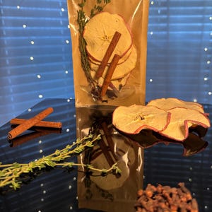May include: A clear, resealable bag filled with dried apple slices, cinnamon sticks, thyme sprigs, and cloves. The bag sits on a reflective surface with additional ingredients scattered around, creating a rustic, natural aesthetic. The ingredients are for a beverage.