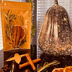 Può includere: Una zucca decorativa in vetro dorato maculato è posta accanto a una busta trasparente piena di fette d'arancia essiccate, bastoncini di cannella, rosmarino e foglie di alloro. Gli oggetti sono su una superficie riflettente.