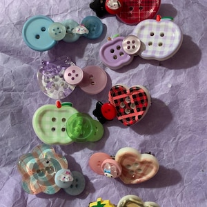 May include: A collection of decorative hair clips featuring various shapes including apples, hearts, and flowers. The clips are adorned with assorted buttons in plaid, gingham, and solid patterns, with small character accents in blue, red, and purple.
