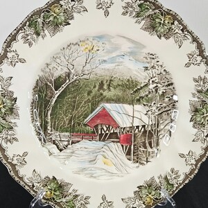 Könnte beinhalten: Ein weißer Keramik-Dinnerteller mit braunem und grünem Blumendekor. Die Mitte des Tellers zeigt eine Winterszene mit einer roten überdachten Brücke, schneebedeckten Hügeln und Bäumen.