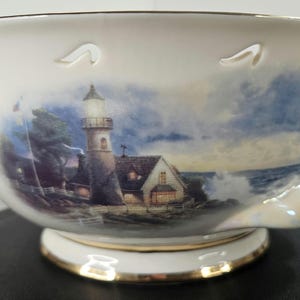 Può includere: Una ciotola in porcellana bianca con un design di faro e paesaggio marino. La ciotola ha un bordo ondulato e un bordo dorato. La ciotola è decorata con un faro, una casa e un mare tempestoso.