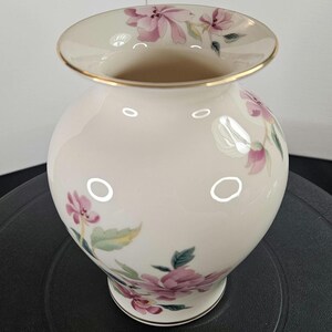 May include: Cream-colored ceramic vase with a round body and a wide, slightly flared rim. The vase is adorned with delicate pink floral patterns and green leaves. A thin gold band accents the rim and base, adding a touch of elegance.