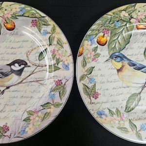 May include: Two white ceramic plates with a floral design and birds. One plate features a blue bird with yellow accents and the other plate features a gray and white bird. Both plates have handwritten text in the background.