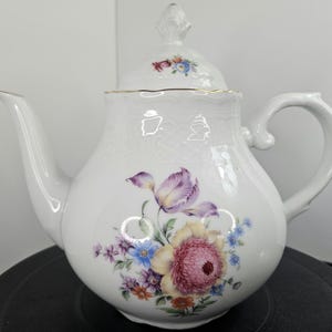 Puede incluir: Tetera de porcelana blanca con un diseño floral. La tetera tiene una tapa con un borde dorado y un asa en el lateral. El diseño floral presenta flores rosas, moradas, amarillas y azules.
