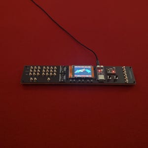 May include: A long, rectangular black electronic circuit board featuring a central color display showing a blue and red graphic, multiple gold-colored knobs, two push buttons, and a black power cable connected to the top of the device.