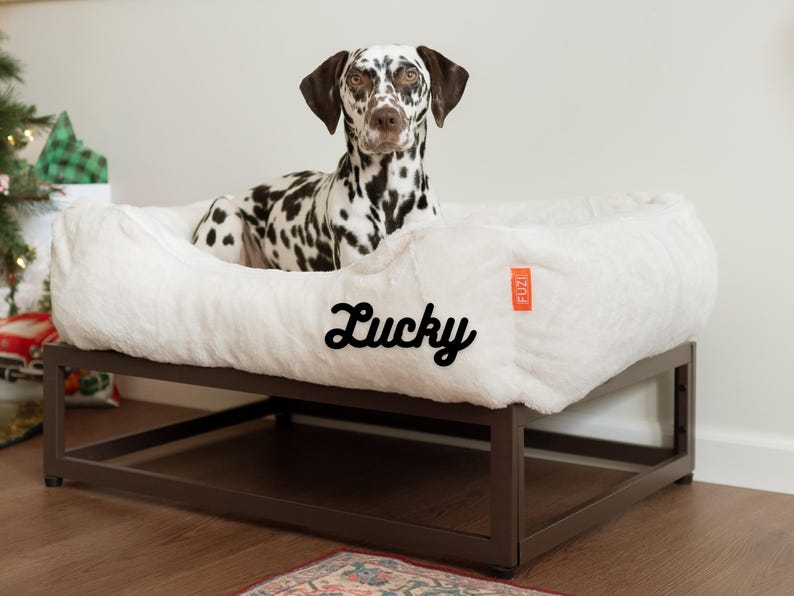 Custom elevated dog bed in soft white faux fur with black metal dog bed frame. Personalized dog bed option with embroidered name. Raised dog bed design functions as a dog window perch or dog window seat, keeping pets comfortable and supported. Orthopedic dog bed for large dogs and small dogs. Washable dog bed with removable faux fur cover for easy cleaning. Modern dog bed doubles as stylish dog crate furniture or pet bed platform. Perfect custom dog bed.