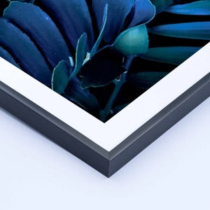 K&ouml;nnte beinhalten: Ein gerahmter Kunstdruck mit einer Nahaufnahme von dunkelblauen und teal-farbenen Bl&auml;ttern. Das Kunstwerk ist in einem wei&szlig;en Passepartout und einem schwarzen Rahmen eingefasst, was eine moderne &Auml;sthetik schafft. Das Bild ist gut beleuchtet und hebt die Texturen und Farben des Laubes hervor.
