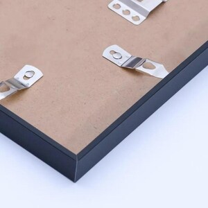 May include: Close-up of a black picture frame with metal hanging brackets attached to the back. The frame has a simple, modern design with a dark finish. The backing is a light brown color.