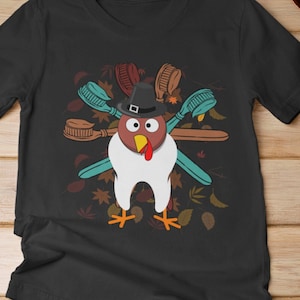 May include: Black V-neck t-shirt with a graphic of a turkey tooth wearing a pilgrim hat and holding toothbrushes. The graphic is surrounded by fall leaves.