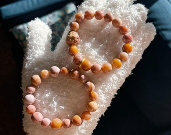 May include: Two bracelets made of pink and orange gemstone beads. The bracelets are on a white fuzzy surface.